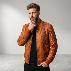 May include: A bomber jacket featuring a vibrant orange and brown snakeskin pattern, a solid orange ribbed collar, cuffs, and hem, and a central front zipper closure. The jacket is worn over a plain black t-shirt and dark-colored trousers.