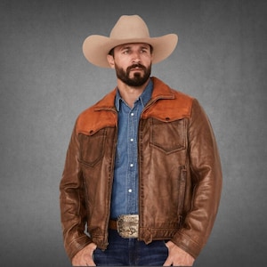 May include: A beige cowboy hat worn with a blue denim button-down shirt and a brown leather jacket featuring a contrasting tan suede yoke across the shoulders. The jacket includes two chest pockets with flaps and a metallic belt buckle at the waist.