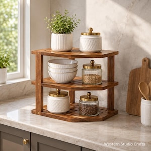 May include: A three-tiered wooden corner shelf featuring a natural brown finish. The top shelf holds a white ribbed planter and a white lidded jar. The middle shelf contains three stacked white bowls and a glass jar with a gold lid. The bottom shelf holds two glass jars with gold lids.