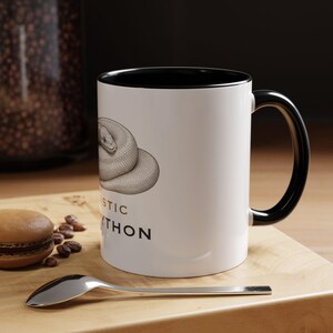 May include: A white ceramic mug with a black interior and handle, featuring a detailed illustration of a coiled snake and the partial text "ISTIC" and "PYTHON" in black and brown lettering printed on the side of the mug's exterior surface.