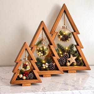 Può includere: Tre cornici triangolari in legno a forma di albero di Natale, ognuna contenente una pallina di vetro trasparente appesa. Le decorazioni includono rami di pino, pigne, piccole sfere dorate, bacche bianche e luci LED calde in ogni elemento.