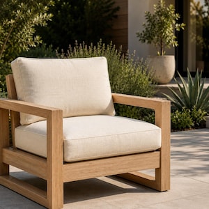 May include: A square outdoor armchair constructed from light-toned wood with a thick, flat-frame base and armrests. The chair features a removable, off-white seat cushion and a matching back cushion, both with a textured fabric finish.