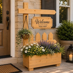 May include: A light brown wooden planter box with a vertical post and a hanging sign that reads Welcome TO OUR HOME. The structure includes a small hanging basket with green foliage and a larger planter filled with white, yellow, and purple flowers.