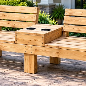 Puede incluir: Un banco de exterior de madera marrón claro con dos zonas de asiento separadas, respaldos de listones y una mesa consola central. La consola central incluye dos portavasos negros circulares integrados en la superficie de madera.