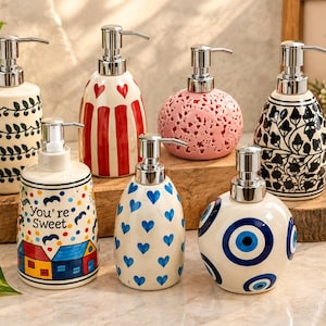 May include: Seven ceramic soap dispensers with silver-tone pumps. Designs include black leaf patterns, red hearts, pink filigree, black floral motifs, a house scene with the text You're Sweet, blue hearts, and blue evil eye symbols on white bases.