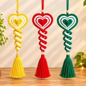 Pode incluir: Três ornamentos suspensos de macramé com um topo em forma de coração com riscas concêntricas, uma secção central em espiral e uma borla comprida na base. Os ornamentos são amarelos, vermelhos e verdes, com detalhes brancos e uma conta de madeira.
