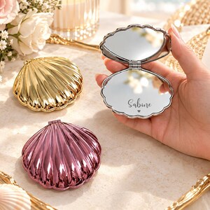 Pode incluir: Três espelhos de bolso em forma de concha com acabamentos metálicos em dourado, ouro rosa e prateado. Um espelho prateado está aberto, revelando uma superfície refletora com o nome Sabine e um pequeno coração impressos em preto.