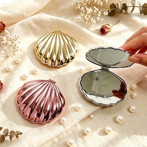 Pode incluir: Três espelhos de bolso em forma de concha com acabamentos metálicos dourados e em ouro rosa. Um espelho está aberto, revelando uma superfície refletora com o nome Regine e um símbolo de coração gravados no vidro. Design canelado e recortado.