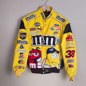 Op de afbeelding: Een felgeel racejack met talloze geborduurde merklogo's, waaronder M&M's, UPS, Ford Racing en Coca-Cola. De voorzijde toont grote M&M's-karakterafbeeldingen, een opstaande kraag, zwarte mouwpanelen en een sluiting met drukknopen.