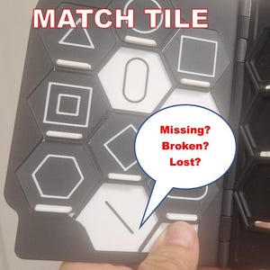 May include: A black plastic game board with hexagonal slots containing white tiles featuring various geometric shapes. Text on the board reads REPLACEMENT MATCH TILE, Solve&Flip, and a speech bubble with the text Missing? Broken? Lost?