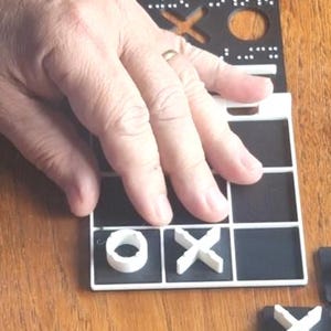 May include: A tactile tic-tac-toe game board featuring a black grid with white borders. The board includes raised white X and O game pieces. The top section of the board displays braille characters and cut-out shapes for tactile identification.