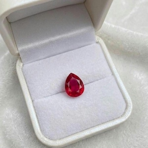 Puede incluir: Una piedra preciosa de rubí rojo facetado en forma de pera presentada dentro de un joyero blanco. La gema presenta un tono carmesí profundo con facetas internas visibles que reflejan la luz, sobre un forro de terciopelo blanco.