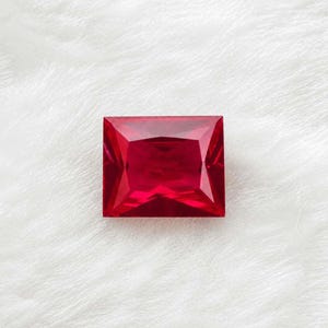 May include: A vibrant, deep red gemstone cut into a rectangular shape with sharp, faceted edges. The stone displays a clear, transparent quality with internal light reflections that create a geometric pattern across its polished surface.