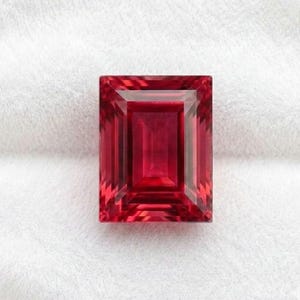 May include: A rectangular gemstone with a vibrant red hue and an emerald cut. The stone features precise, symmetrical facets that create a clear, reflective surface with deep crimson tones throughout the center and lighter red edges.