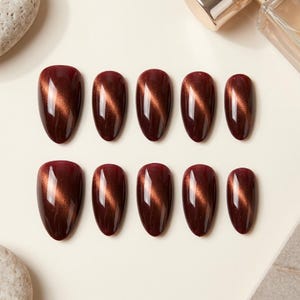 Peut inclure: Un ensemble de dix faux ongles en forme d'amande d'une couleur bordeaux profonde et brillante. Chaque ongle présente un effet magnétique œil-de-chat scintillant qui crée une ligne de lumière verticale au centre de la surface rouge sombre.