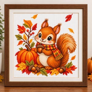 May include: A framed cross-stitch embroidery featuring a brown squirrel wearing a plaid scarf, holding an acorn. The squirrel is surrounded by an orange pumpkin, red and yellow autumn leaves, and scattered acorns on a white fabric background.