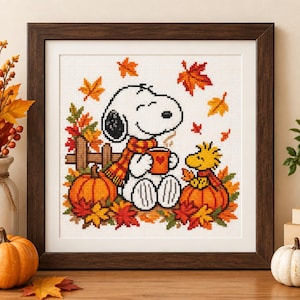 Puede incluir: Un bordado en punto de cruz enmarcado que muestra a Snoopy y Woodstock rodeados de hojas de otoño y calabazas. Snoopy lleva una bufanda de cuadros naranja y amarilla mientras sostiene una taza con un corazón. La escena incluye una valla de madera.