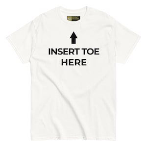 Puede incluir: Una camiseta blanca de manga corta con un gráfico de flecha negra que apunta hacia arriba sobre el texto negro en negrita que dice INSERT TOE HERE. La prenda tiene un cuello redondo estándar y una pequeña etiqueta rectangular en el interior.