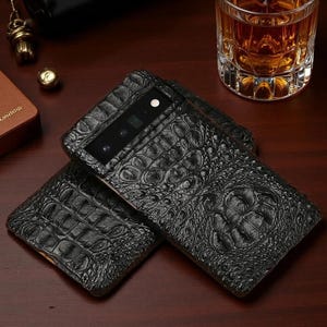Peut inclure: Une coque de smartphone noire avec un motif texturé en peau de crocodile. La coque présente une découpe rectangulaire en haut pour le bloc photo, comprenant plusieurs objectifs circulaires et un flash. La surface a une finition gaufrée brillante.