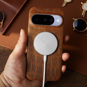 Peut inclure: Une coque de smartphone marron avec un motif texturé en peau de crocodile, dotée d'une découpe horizontale pour l'appareil photo en haut et d'un chargeur magnétique sans fil blanc circulaire fixé au centre avec un câble blanc vers le bas.