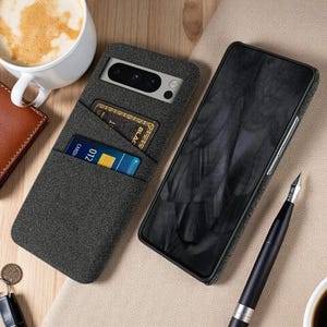 May include: A dark gray fabric-covered smartphone case featuring a rear card slot containing two credit cards. The case is shown from both the back, displaying the camera cutout, and the front, showing the black screen of the mobile device.