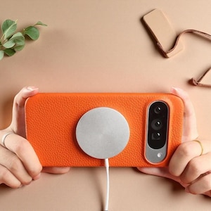 Op de afbeelding: Een feloranje smartphonehoesje met een getextureerde afwerking en een zilverkleurige, ronde magnetische draadloze oplader in het midden. Het hoesje heeft een uitsparing voor de cameramodule met drie lenzen en een flitser in een zilveren frame.