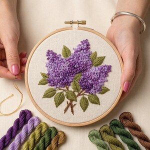 May include: A circular wooden embroidery hoop containing a fabric panel with a hand-stitched design of purple lilac flowers and green leaves. The embroidery features textured French knots and satin stitches, accompanied by various coloured embroidery floss.
