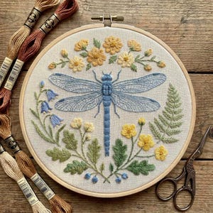 Puede incluir: Un bastidor de bordado circular de madera que contiene un panel de tela con un diseño bordado a mano de una gran libélula azul centrada entre flores amarillas, hojas verdes, flores azules en forma de campana y un helecho.