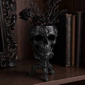 Peut inclure: Un pot de fleurs en forme de crâne noir avec des sculptures florales complexes et une base sur pied. Le pot contient un arrangement sombre composé d'une rose noire unique et de branches fines et nues, formant un objet décoratif détaillé.