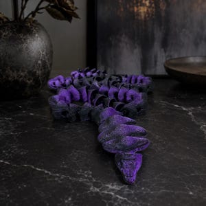 May include: A flexible, articulated snake figurine featuring a gradient colour pattern that transitions from deep black to vibrant purple. The segmented body is composed of multiple interlocking pieces, creating a realistic, winding, and serpentine form.