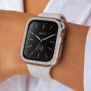 Pode incluir: Um smartwatch com pulseira de silicone branca e uma caixa protetora prateada com uma borda de pequenos cristais transparentes. O mostrador digital preto exibe a data SAT 23 e uma complicação circular com o número 74.