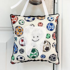 May include: A square fabric pillow featuring a pattern of colorful soccer balls on an off-white background. A white felt tooth shape with a stitched smiling face is centered on the front. The pillow hangs from a white ribbon tied to a door knob.