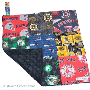 May include: A patchwork baby blanket featuring logos of Boston sports teams: Patriots, Bruins, Celtics, and Red Sox. The blanket has a black, textured backing and a loop with a Bruins logo. The blanket is made of various fabrics.