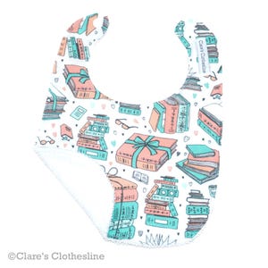 May include: A white baby bib with a pattern of stacked books in shades of coral and teal, tied with teal bows. The bib features a soft, absorbent backing and a curved shape for comfort. The bib is decorated with hearts and glasses.
