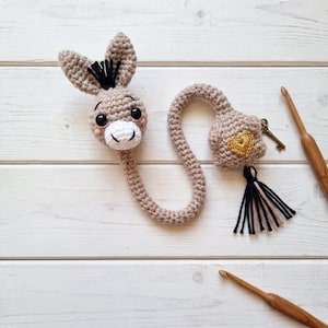 Puede incluir: Un marcapáginas de ganchillo beige con forma de burro, cabeza con pelo negro, ojos oscuros y hocico blanco. El cuerpo largo se conecta a una pieza cuadrada decorada con un corazón bordado en oro, una pequeña llave de metal y una borla bicolor.