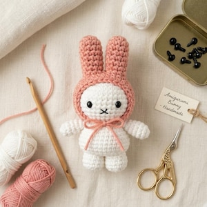 Può includere: Un coniglietto amigurumi all'uncinetto bianco che indossa un cappuccio rosa polvere con un fiocco abbinato. Il coniglietto ha occhi a perla neri e una bocca ricamata. Accanto ci sono un uncinetto in legno, filato rosa e bianco, forbici dorate e un'etichetta con scritto Amigurumi Bunny - Handmade.