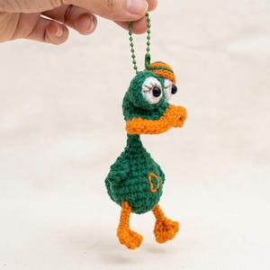 Puede incluir: Un pequeño llavero de pato tejido a mano en crochet, con cuerpo y cabeza de color verde oscuro, pico, patas y una pequeña gorra de color naranja brillante. El pato tiene grandes ojos blancos con pupilas negras y una letra D naranja bordada.