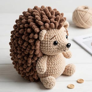May include: A crocheted hedgehog plush toy featuring a textured brown back made of looped yarn and a smooth beige face, belly, and limbs. The toy has two small black plastic eyes and a black plastic nose, with stitched eyebrows above the eyes.
