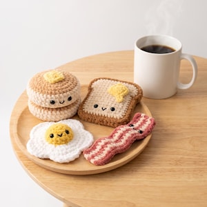 Puede incluir: Un conjunto de cuatro artículos de desayuno tejidos a ganchillo que incluye una pila de dos tortitas color canela con un trozo de mantequilla amarillo, una tostada marrón con mantequilla, un huevo frito blanco con yema amarilla y una tira de tocino rosa y blanca.
