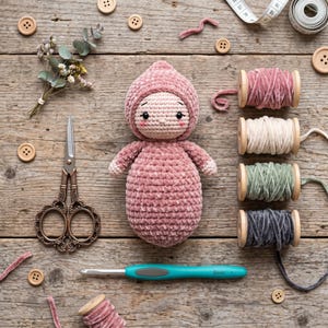May include: A crocheted doll in a dusty pink hooded outfit with embroidered facial features, surrounded by four spools of velvet ribbon, a pair of ornate metal scissors, a 5mm crochet hook, wooden buttons, and a small dried flower bouquet.