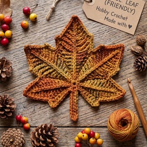 May include: A crocheted maple leaf in variegated autumn colors of orange, yellow, and brown. The leaf features a textured stitch pattern and a central stem. A wooden crochet hook, a ball of yarn, pinecones, and small berries surround the leaf.