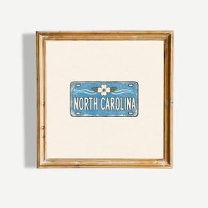 May include: A square wooden frame containing a print of a light blue number plate with a white dogwood flower graphic and the text NORTH CAROLINA in white capital letters, centred on a plain off-white background within the frame.