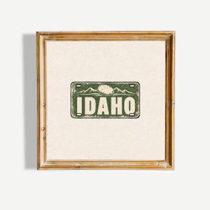 Puede incluir: Una impresión cuadrada enmarcada que presenta un gráfico de matrícula rectangular verde con una textura desgastada. El gráfico muestra la palabra IDAHO en letras blancas grandes, coronada con una ilustración de patata y montañas.