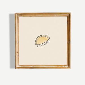 May include: A square framed wall art piece featuring a light cream background with a centred, hand-drawn illustration of a yellow pierogi. Below the pierogi, the word PITTSBURGH is written in black, simple, uppercase capital letters.