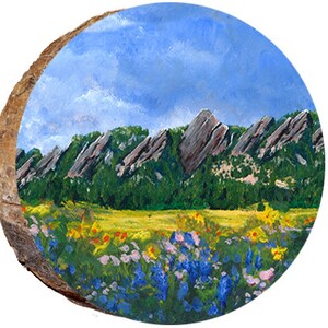 May include: A painting on a wood slice depicting a landscape with a mountain range in the background and a field of wildflowers in the foreground. The sky is blue with white clouds. The mountains are a mix of brown and green, and the wildflowers are a variety of colors, including blue, purple, yellow, and pink.