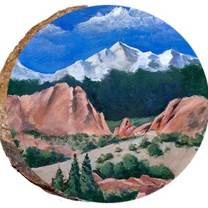 May include: A round, hand-painted landscape scene on a wood slice. The artwork depicts a mountain range with snow-capped peaks under a blue sky with clouds. Red rock formations and green trees fill the foreground.