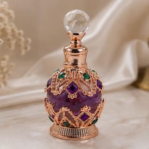 May include: A decorative perfume bottle featuring a round purple glass body encased in an ornate rose gold metal filigree frame. The frame is accented with small green and purple faceted stones. A clear crystal topper sits on the bottle neck.