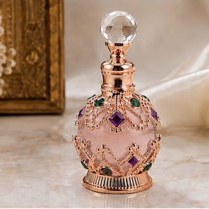 May include: A round glass perfume bottle with a pale pink body, encased in an ornate rose gold metal filigree frame. The bottle features a clear faceted crystal stopper and is decorated with small, embedded purple diamond-shaped and green oval-shaped gems.