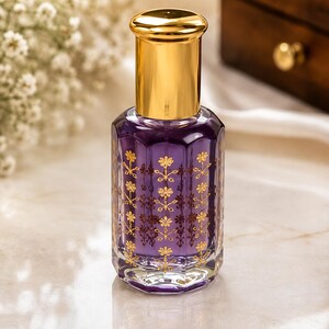 May include: A glass perfume bottle with a purple liquid interior and a gold-colored cap. The bottle features a hexagonal shape and is decorated with a repeating gold floral pattern printed on the exterior glass surface of the container.