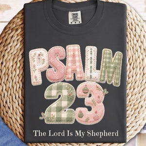 May include: A dark gray t-shirt featuring the text PSALM 23 in a patchwork-style font with pink, green, and white floral and gingham patterns. Below the large text, the phrase The Lord Is My Shepherd is printed in white. The shirt has a crew neck.